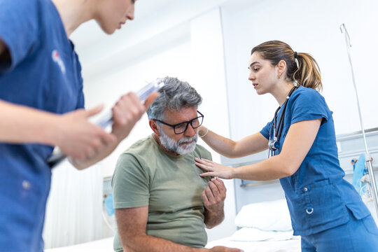 Female doctor examining an elderly male patient sitting on a hospital bed complaining of chest pain, while another female doctor holds medical documents in a modern clinical room.