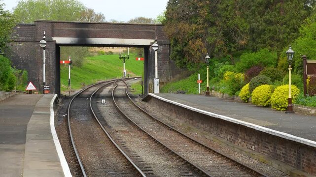 A wide shot of a quiet historic train station platform featuring parallel tracks leading under a brick bridge. Suitable for travel and transport documentaries.