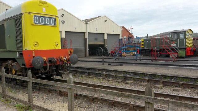 A static shot of classic British diesel trains parked outside maintenance sheds at a heritage railway station. Suitable for transport history and industrial documentaries.