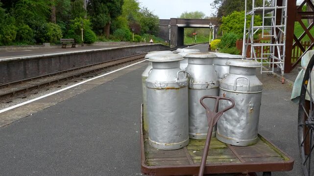 A collection of traditional metal milk churns sits on a wooden trolley at a quiet rural railway station. Suitable for historical documentaries and heritage travel content.