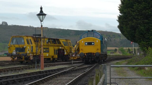 A classic blue and yellow diesel locomotive passes a yellow track maintenance vehicle on parallel tracks in a rural English railway yard.