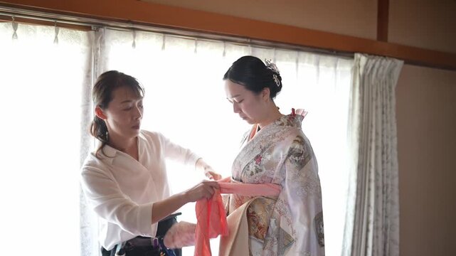 Soft light filters through wooden-framed curtains. Woman in a kimono stands calmly, facing away. Another assists her, adjusting the garment gently. The scene conveys quiet, respectful care