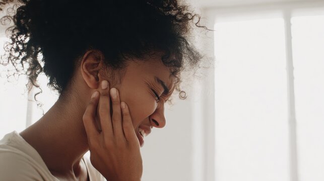 Woman holds her face with pain while standing in a bright room during the day