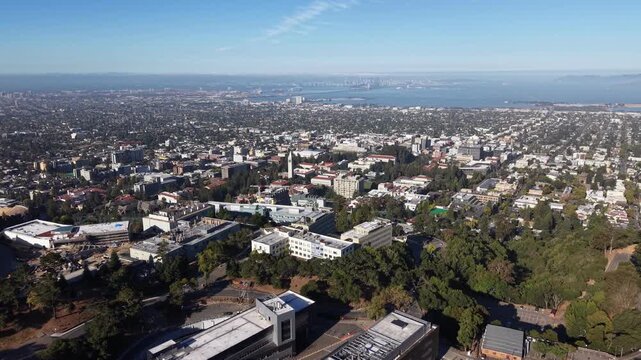 Aerial view showing the University of California, Berkeley campus with the San Francisco Bay, Oakland, and the Bay Bridge visible in the distance on a clear and sunny day. Rising drone shot