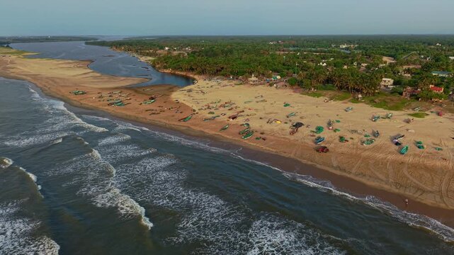 Aerial drone shot of Bay of Bengal waves rolling onto Chennai beach where fishing boats line the sandy shore beside backwaters and coastal vegetation in Tamil Nadu, India
