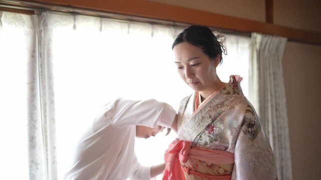 Soft light fills a quiet Japanese room. Woman adjusts a kimono with care and focus. Another woman helps with the sash and folds. Their expressions show gentle concentration