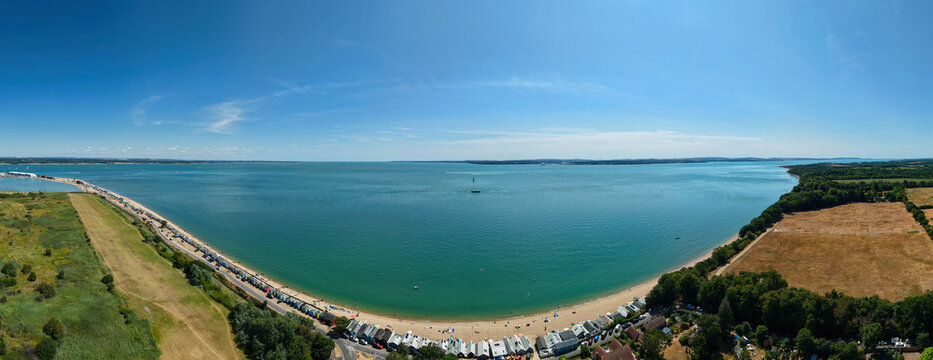 Aerial view of Calshot Beach with its iconic row of beach huts along the coastline, surrounded by the blue sea and green fields in Calshot, England, United Kingdom.