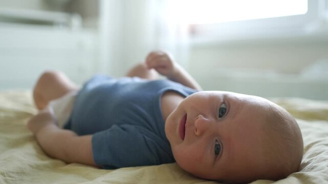 Sweet newborn baby boy lying on his back in a sunlit bedroom, experiencing a whirlwind of emotions from happiness to sadness, his expressions changing from smiling to crying