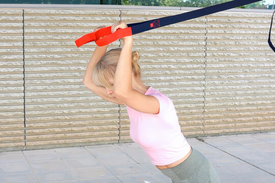 Young woman performing suspension trainer chest press outdoors beside textured wall, wearing light pink top and gray leggings, with focused effort, strong posture, and bright sunlight
