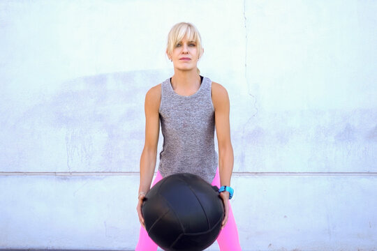 Athletic woman holding heavy medicine ball during strength exercise against plain white wall, focused posture, sleeveless gray workout top, bright pink leggings, full body fitness portrait
