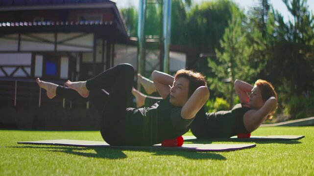 Active senior women practicing slow-motion Pilates together on exercise mats outdoors, performing controlled core strengthening stretches on sunny green grass