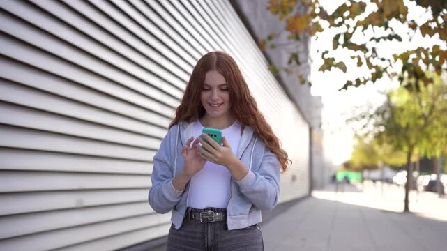 A Young Woman Delightfully Enjoying Her Smartphone While Leisurely Walking Outdoors in the City