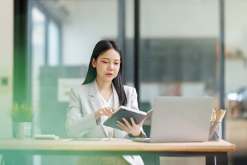 Asian businesswoman reviewing agenda in notebook, planning strategy in modern office, using laptop...