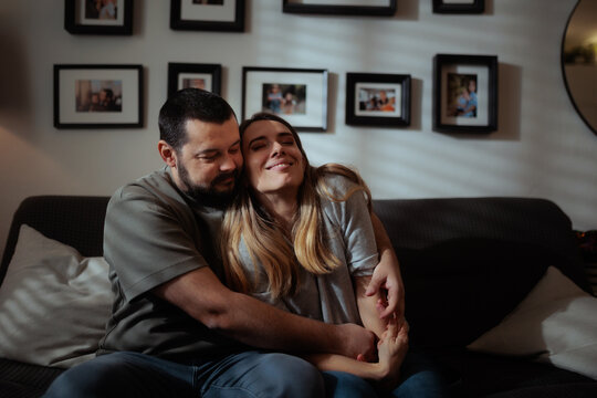 Happy Wife Smiling While Embracing Her Loving Husband On Sofa. Cheerful married couple enjoys romantic affectionate moment together hugging on cozy living room couch
