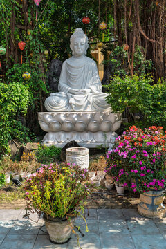 White stone Buddha statue sitting in lotus position in the courtyard of the Phap Bao Pagoda (Chua Phap Bao) surrounded by hanging lanterns and flowers in Hoi An, Vietnam.