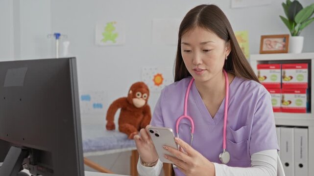 Woman doctor in purple scrubs taps smartphone while stethoscope rests around her neck in a pediatric clinic building with a stuffed monkey on the exam table; focused care.