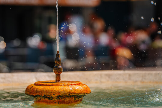Close-up of a decorative fountain with water spouting from the center, surrounded by blurred figures in a lively outdoor setting in Bergamo