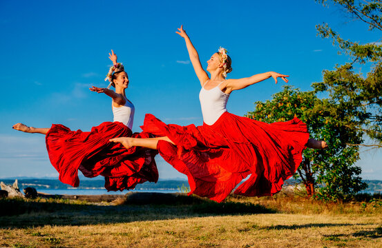 Two white women jump gracefully outdoors wearing red skirts and flower
