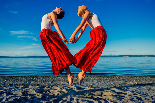 Two white women performing partner acrobatics yoga jumps on a beach