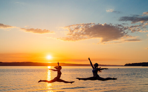 Two white women performing split leaps over water at sunset in WA, USA