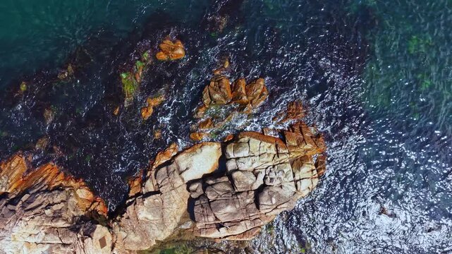 Bright sunlight shines on the ocean, highlighting the rocky shore. Waves crash against the stones, creating splashes. The scene shows natural patterns of water and rock.