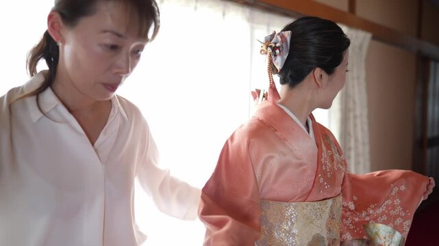 Soft light bathes a traditional room. Woman adjusts a kimono's sash with gentle care. Another woman stands, poised, wearing ornate attire. They share quiet intimacy near a window