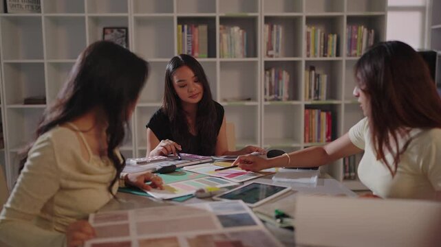 Close view of three designers examining bright paint chips and sketches together as one points a pen toward color coded swatch cards on the table