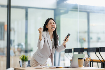 Fototapeta premium Young asian businesswoman cheering, raising a fist, and holding a phone, celebrating an achievement in a modern office