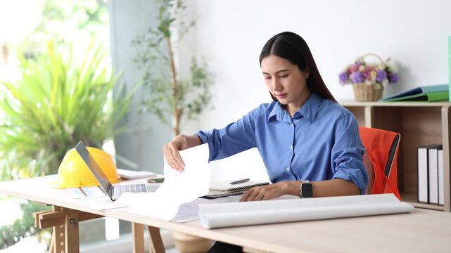 Young female asian civil engineer or architect checking a building blueprint, professional woman at work in her office inspecting a project and working on her laptop
