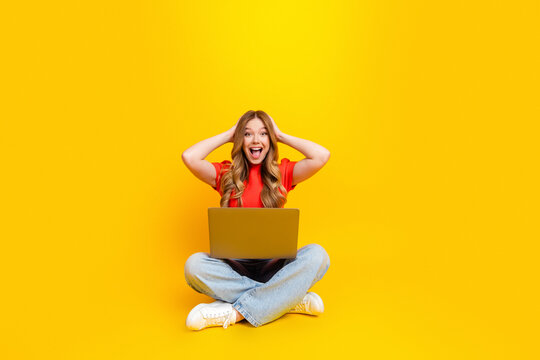 Excited young woman sitting cross-legged with laptop on bright yellow background celebrating good news or online success