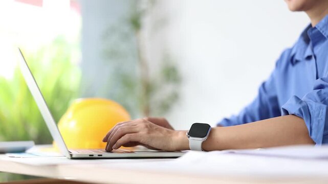 Professional architect typing on laptop in the office while working on building plan blueprints. Female engineer developing a construction project, wearing a smartwatch
