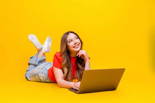 Young woman lying on a bright yellow background with laptop, smiling and working or studying while daydreaming and creating ideas