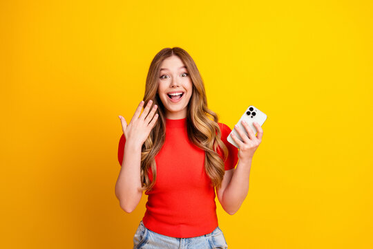 Happy young woman surprised and excited holding smartphone against a bright yellow background, casual red top and long hair, joyful reaction and modern mobile communication portrait