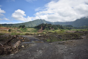 Dirt road winds through rugged terrain. Mist drapes distant, green mountain peaks. Clouds drift softly above the hills. Vast land reveals signs of recent clearing © Fajar
