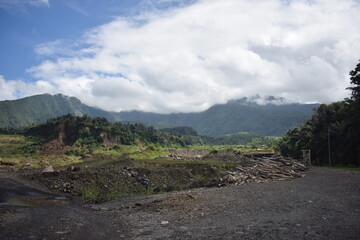 Dirt road winds through rugged terrain. Mist drapes distant, green mountain peaks. Clouds drift softly above the hills. Vast land reveals signs of recent clearing © Fajar