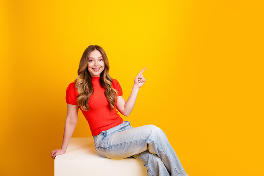 Smiling young woman in red top sitting on a cube and pointing to empty space on bright yellow background