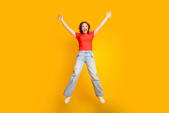 Happy young woman jumping with arms raised on bright yellow background expressing excitement and energy in a casual red top and wide-leg jeans