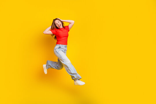 Young woman jumping joyfully against a bright yellow background in a red top and jeans, expressing happiness, energy and casual lifestyle for advertising and social media