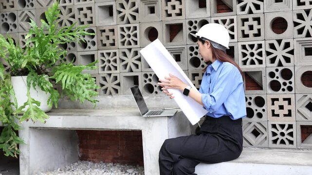 Professional asian female civil engineer with a hard hat checking blueprints and talking on the smartphone while working outdoors on a laptop at a construction site
