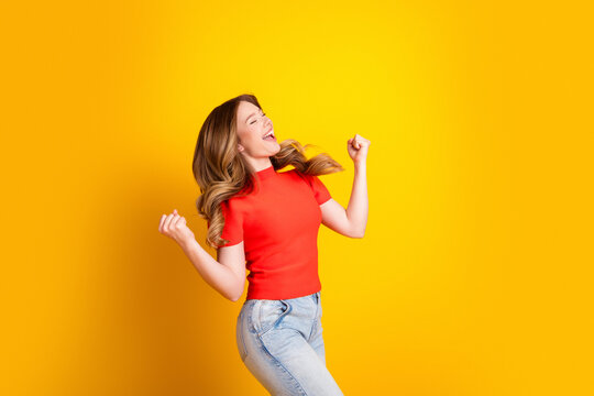 Joyful young woman celebrating with raised fists against a bright yellow background in a red shirt and jeans, energetic portrait of happiness and victory