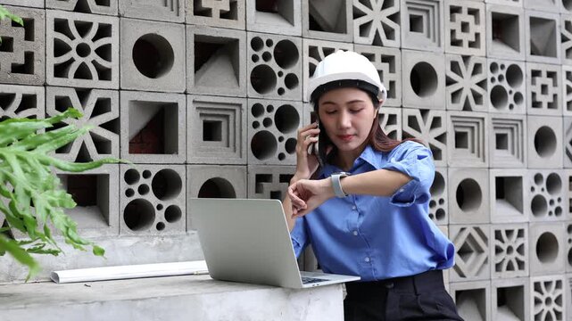 Female civil engineer wearing a hard hat working on a laptop computer and talking on the smartphone while multitasking on a construction project in an outdoor modern setting