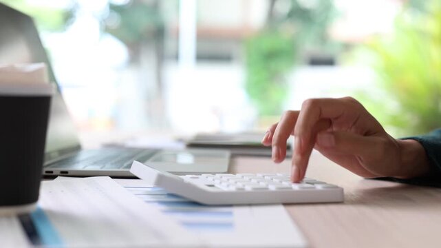 Businesswoman calculating company finances and expenses with a calculator at her desk, analyzing profit and revenue data from financial reports for investment planning