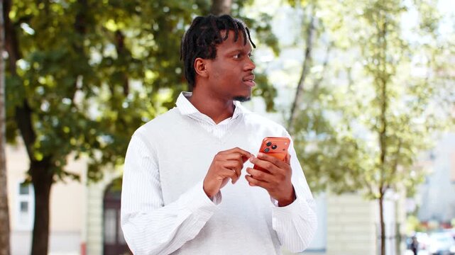 African American young man checking smartphone walking in city street with calm mood during park travel. Black guy tourist looking around outdoors using mobile app for map route search in urban trip.
