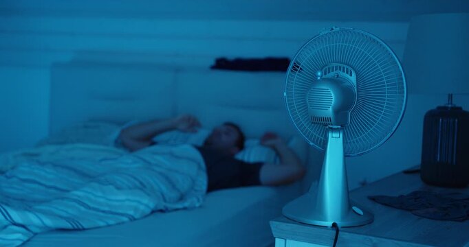 A man struggles to fall asleep on a hot summer night despite using an electric fan.
