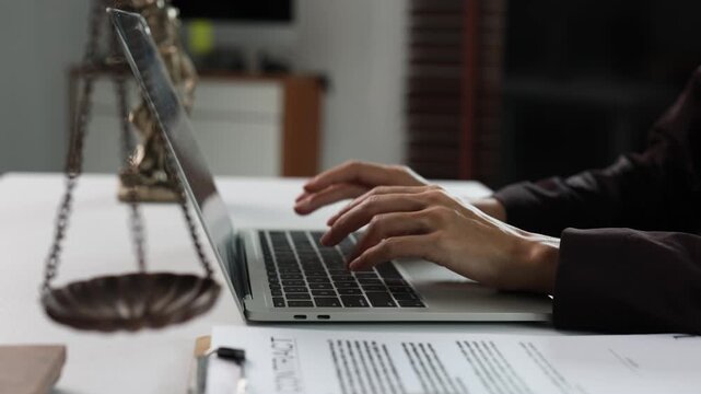 Lawyer typing on a laptop with a contract and scales of justice on the desk, female attorney working in a law firm office preparing legal documents for court