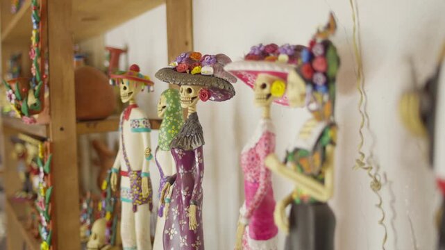 Decorative Catrina figurines displayed on shelves inside artisan workshop, pastel white and pink purples clothes, shallow depth of field