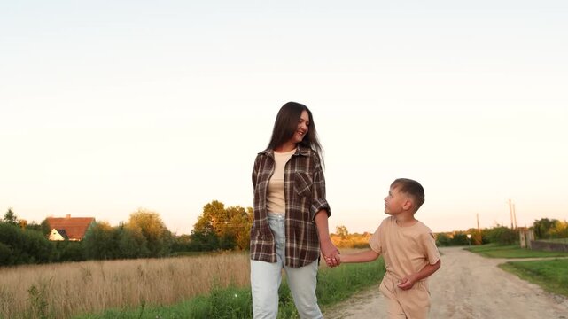Happy mother and child walk hand in hand along sandy village path before sunset. Slow motion captures smiling faces and warm connection during peaceful evening.
