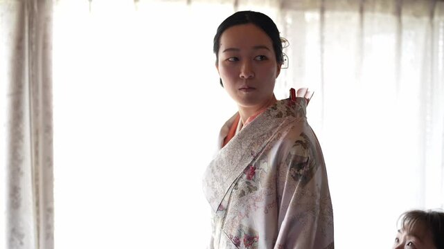 Soft light filters through sheer curtains. Woman adjusts the other's kimono with care. The second woman stands still, eyes closed, calm. The quiet moment captures a tender, personal ritual