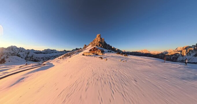 Distorted wide angle drone view of refuge with Ra Gusela rugged snow covered mountain peak illuminated by golden sunset light at Giau Pass in Italy. Aerial forward