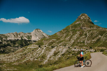 Fototapeta premium Female cyclist with gravel bike stopping on the mountain road at Sedlo Pass in Durmitor National Park, Montenegro. Stunning alpine scenery surrounds the scenic cycling route